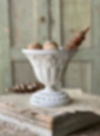 A white distressed French urn with intricate details, styled with rustic wooden beads and decor, sitting on vintage papers