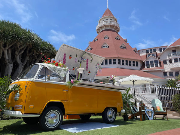 Vintage yellow 1972 VW bus mobile bar set up outside the Hotel del Coronado in San Diego, decorated with flowers, umbrellas, and lounge seating for an outdoor event.