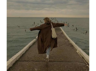Person in a brown coat runs on a pier with arms outstretched, surrounded by flying birds. Overcast sky, ocean backdrop, evokes freedom.