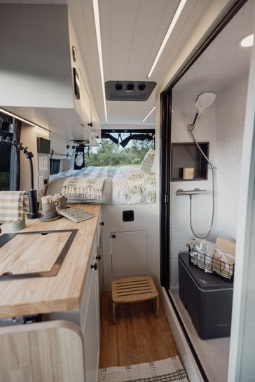 Camper van interior view showing kitchen, shower, and bed layout.