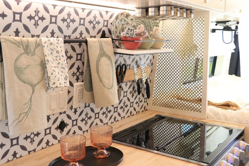 Camper van kitchen backsplash with hanging towels, knives, and open shelf of dishes.