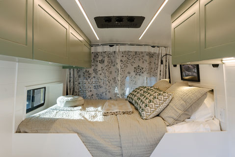 Camper van bed with floral curtains and green overhead cabinets.