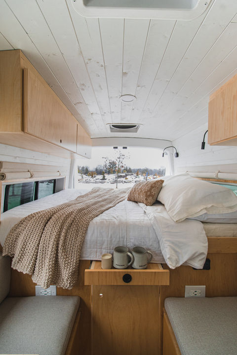 Custom camper van bed area with wood cabinetry, beige knit blanket, and soft white bedding.