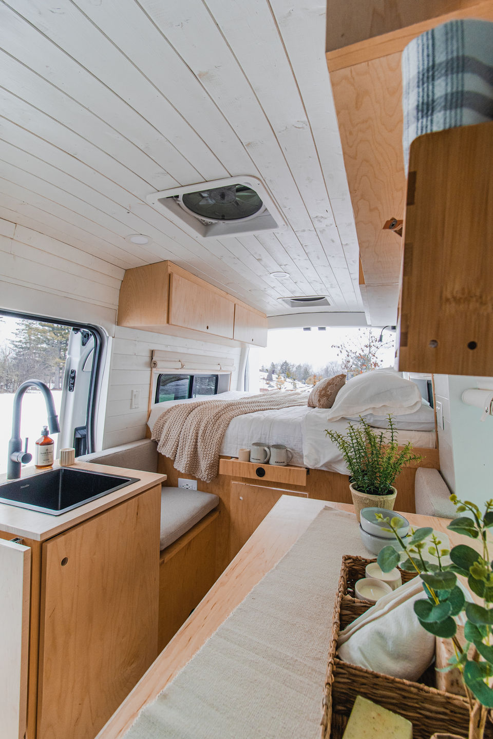 Wide view of custom camper van interior showing kitchen and bed area with light wood finish.