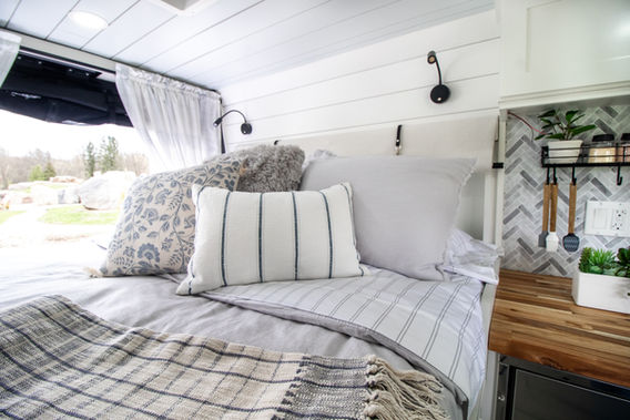 Camper van bed with striped pillows and gray blanket beside window.