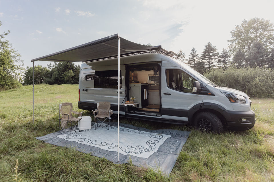 Camper van parked on grass with outdoor rug and awning extended.