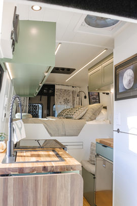 Camper van interior showing kitchen and bed area with sage cabinets and white walls.