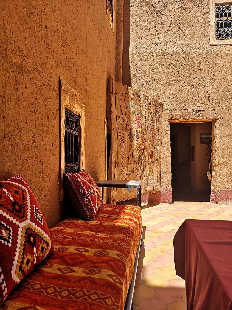 Sunlit rustic courtyard with red patterned cushions, wooden table, painted door, and open doorway in mud-brick walls.