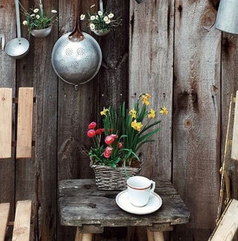 Two wooden chairs face a rustic wooden wall adorned with hanging kitchen utensils. A small table holds a teacup and a flower basket. Cozy setting.