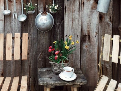 Two wooden chairs face a rustic wooden wall adorned with hanging kitchen utensils. A small table holds a teacup and a flower basket. Cozy setting.