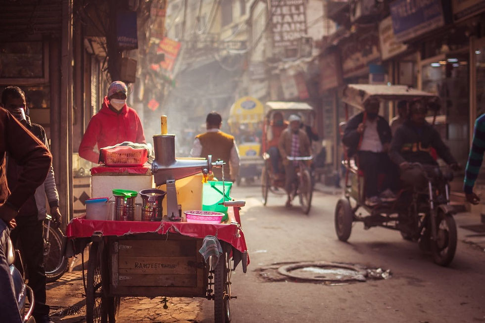 Masked street vendor in red hoodie serves drinks from a cart in a hazy crowded alley with rickshaws
