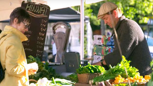 Man and woman at an outdoor market stand. Man in cap weighs produce beside baskets of greens and flowers. Price sign reads "Hinnat".