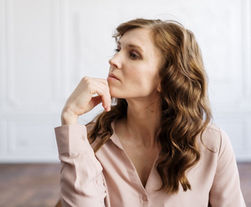 Woman in a pink blouse, sitting in a bright room, looking thoughtful with her chin on her hand. Curly hair, serene atmosphere.
