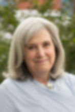 Elderly woman with gray hair smiles gently, wearing a blue blouse in a garden setting with blurred greenery in the background.