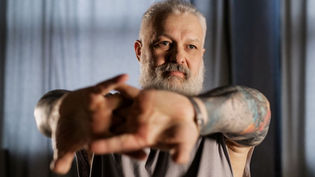 Young looking middle-aged bearded man with tattoos stretches arms forward, appearing focused. Soft lighting, blurred dark curtains in the background.