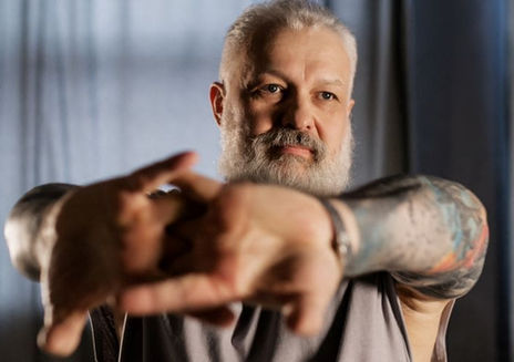 Young looking middle-aged bearded man with tattoos stretches arms forward, appearing focused. Soft lighting, blurred dark curtains in the background.