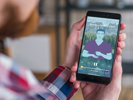 Person holding a smartphone showing a meditation app with "Release General Pain" on screen. Background features a forest image and control icons.
