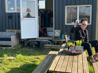 Vrouw in het zwart, glimlachend en heffend het glas op een buitentafel met een kat en bloemen. Een piepklein huisje op de achtergrond, gras op de voorgrond.