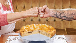 Elderly woman and tattooed arm fist bump over partially eaten pie on lace-covered table. Cozy setting with brown tufted background.