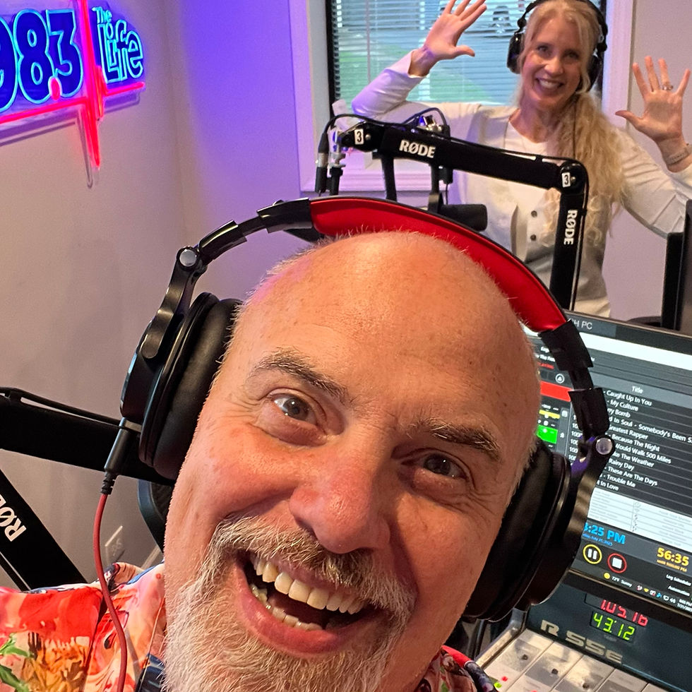 Marty Jalove, Master Happiness and Tiffany smiling in a radio studio. Man in floral shirt, woman raising hands. Neon sign reads "98.3 The Life." Playful mood.