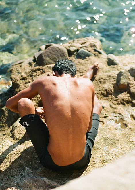 A person sits on rocks by a body of water. From the back, the person is shirtless and sun reflects off their skin.