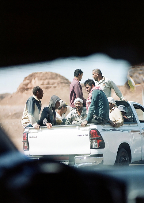 An image captures a candid scene of several men riding in the back of a white pickup truck, partially obscured by a dark object on the left, foreground.