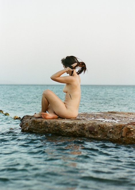A nude woman with dark hair is sitting on a rock in the ocean facing right. She has her hand on her head securing her hair so that it is up in a high ponytail.