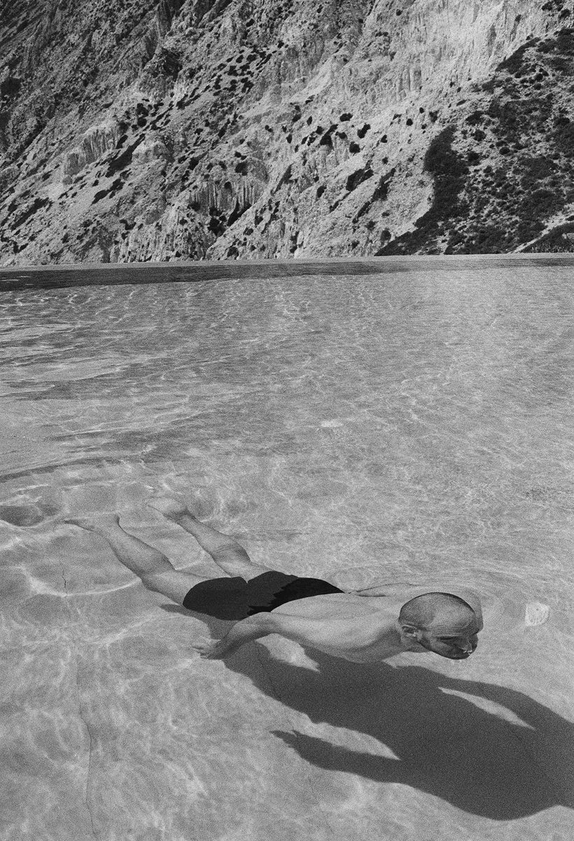 The black-and-white photo captures a swimmer in clear water, with a large mountain behind them.