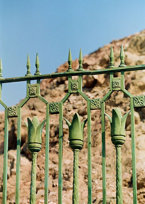 The image shows a close-up of a green iron gate in front of a rocky hill. The gate features decorative elements including tulip-shaped ends and sharp, pointy tops.