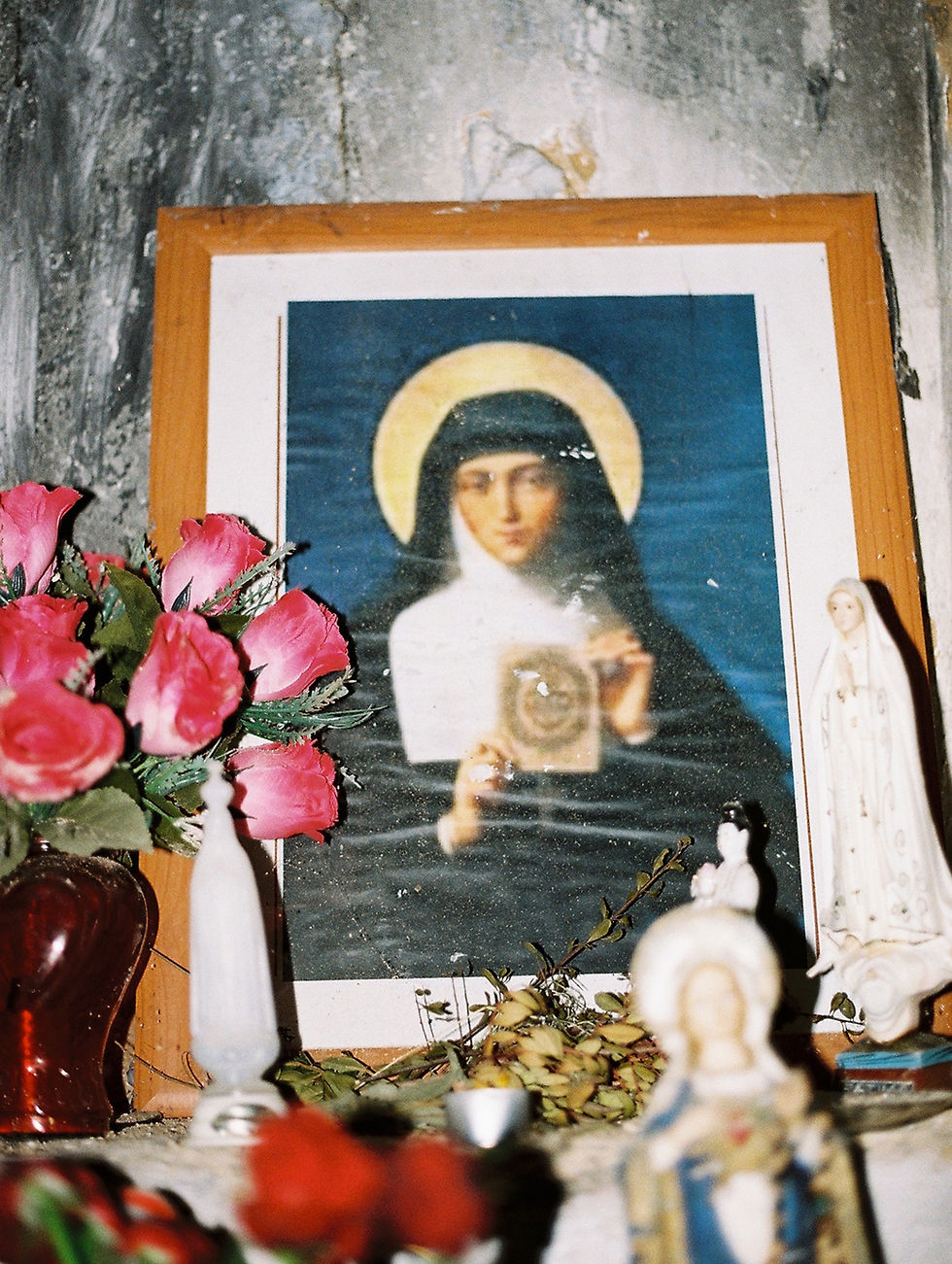 An indoor shrine, possibly of Saint Teresa, is shown in a medium shot. In the center is a framed portrait of a woman in a nun's habit, an ornate halo, and a blue background, holding a gold object.