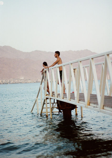 A photo captures a couple on a pier. A man stands shirtless, looking out toward a hazy mountain range.