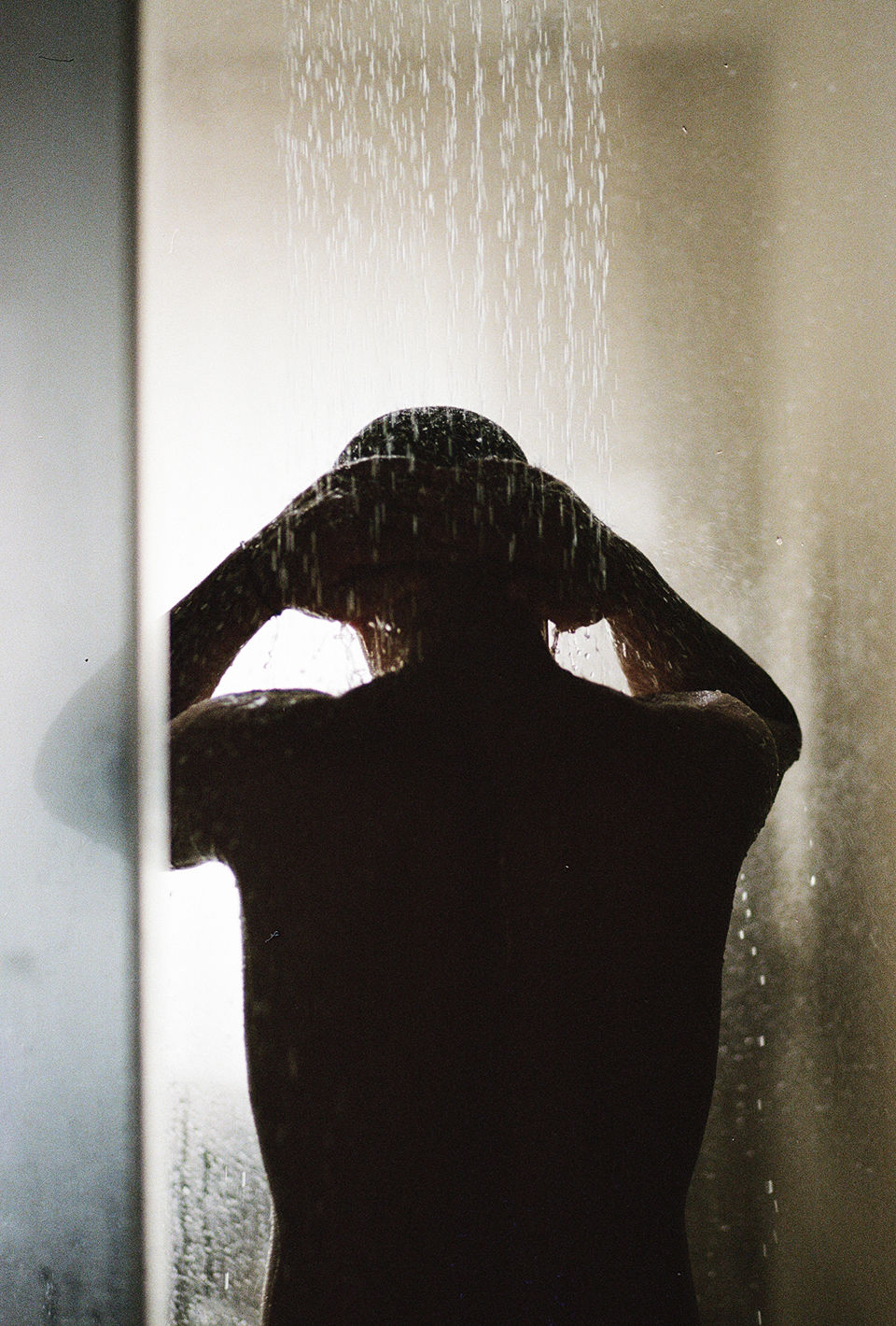A person stands with their back turned, taking a shower. The person is in silhouette, with water pouring down over their head and hands.
