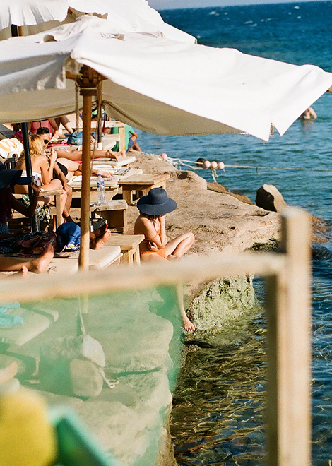 A sunny beach resort scene with a woman in a hat in the foreground. She sits cross-legged on a concrete ledge, wearing an orange swimsuit. 