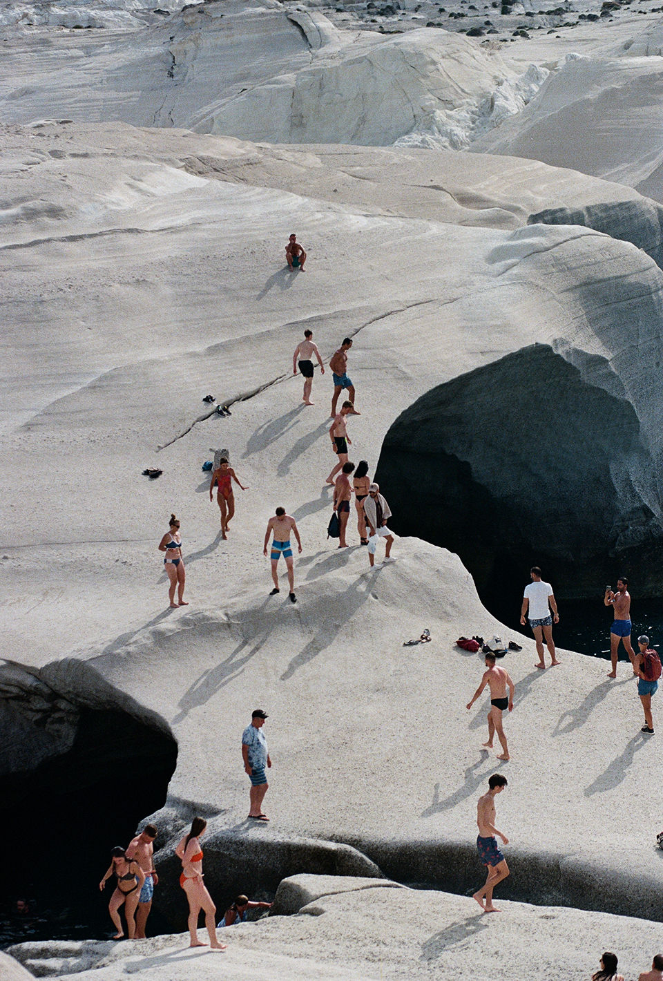 People in swimwear populate a light-filled outdoor scene. They are on a landscape of pale white, smooth rock formations, likely near a body of water.