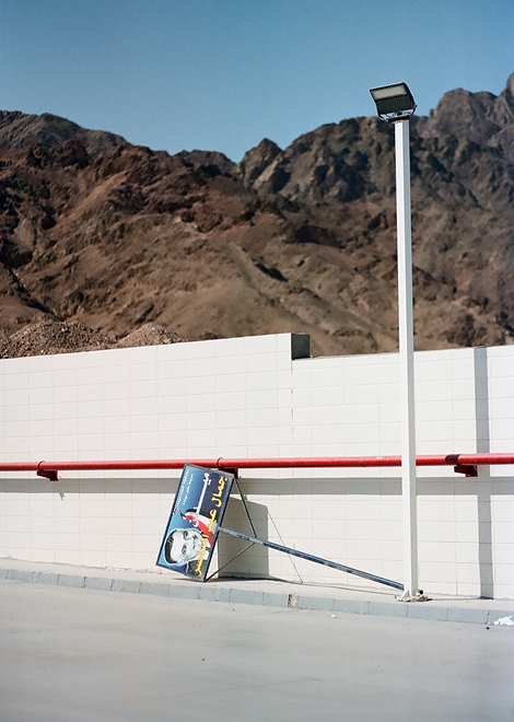 A eye-level shot depicts a street with a sign leaning against a white wall and a mountain range in the background under a clear sky.
