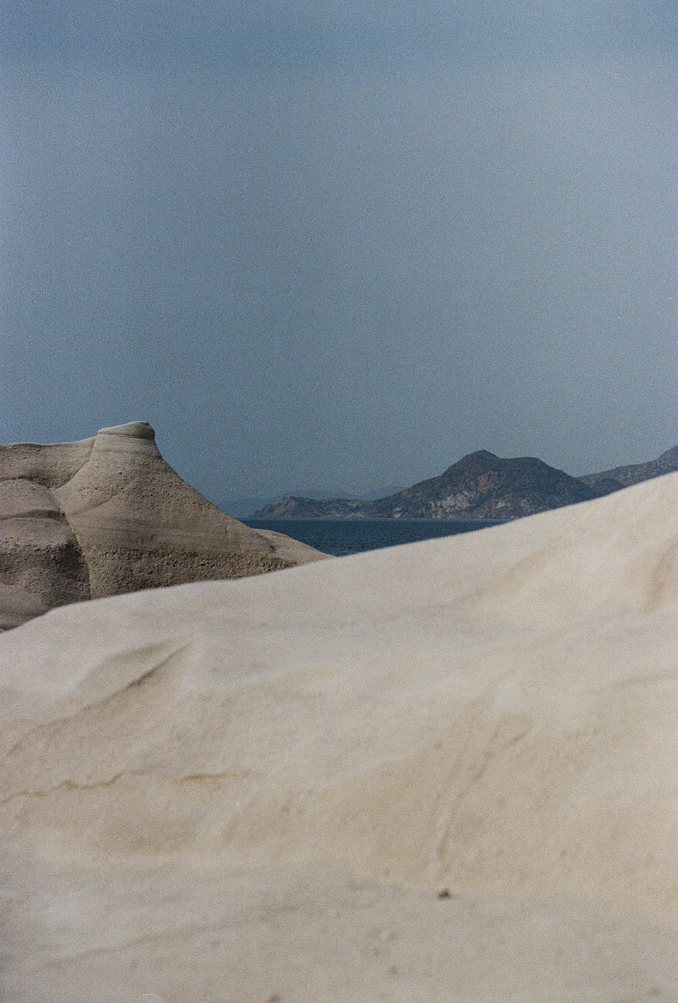 A scenic photograph captures a grayscale landscape under a pale blue sky. In the foreground, a windswept sand dune stretches, while in the background are hills and the ocean in between them.