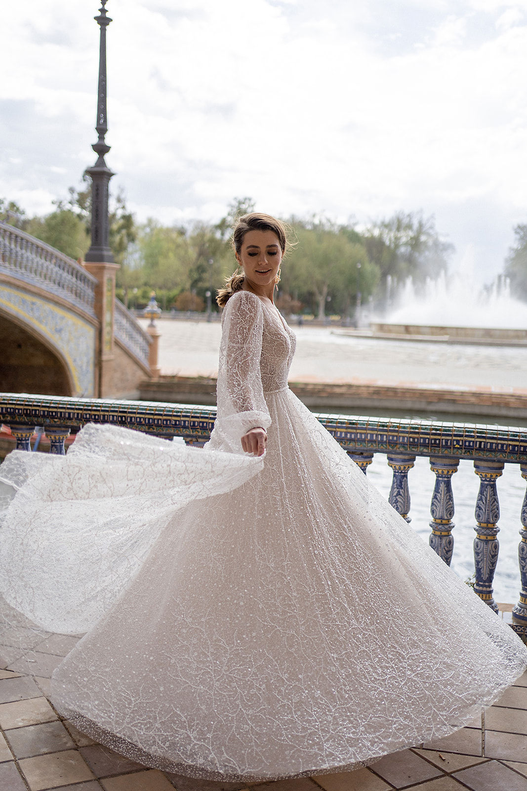 Suri wedding dress by Dama Couture – A-line gown with corset bodice, floral appliqué, glitter tulle, sheer sleeves, and deep