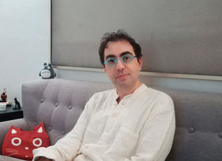 A man in a white shirt and round, sky blue glasses sits on a grey couch. Next to him are a red pillow in the shape of a cat face and a small Totoro figurine