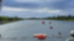 Swimmers in a canal race, overseen by people in kayaks. Red buoys mark lanes. Lush green banks and a cloudy sky create a vibrant scene.