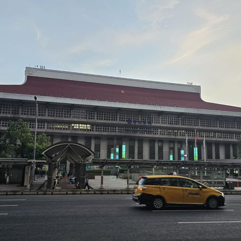 A large, modern building with a red roof and grid windows at dusk. Yellow taxi on the road in front, with a clear sky overhead.