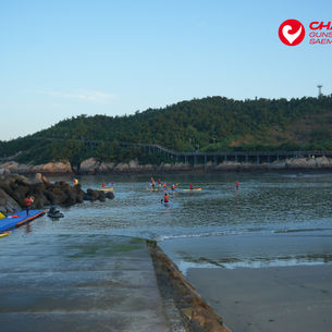 Paddleboarders and swimmers in a calm coastal bay near a green hill; red CHALLENGE GUNSAN SAEMANGEUM logo at top right