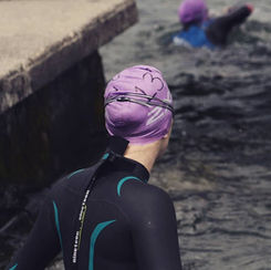 A person in a black wetsuit and purple swim cap stands by water, looking at a swimmer in similar gear. Concrete dock and dark waves visible.