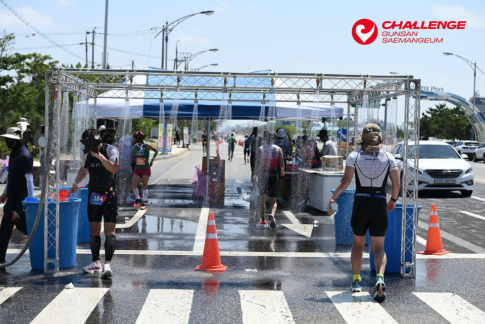Runners pass through a water spray station at the Challenge Gunsan Saemangeum race on a sunny street, with cones and tents.