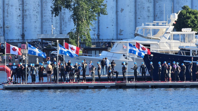 Athletes lined up on a pontoon. Wearing wetsuits and blue swim caps. Quebec and Canadian flags on the pontoon
