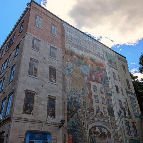 Mural of old buildings on the side of a building. The mural has people looking out of windows and wandering infront of the old building