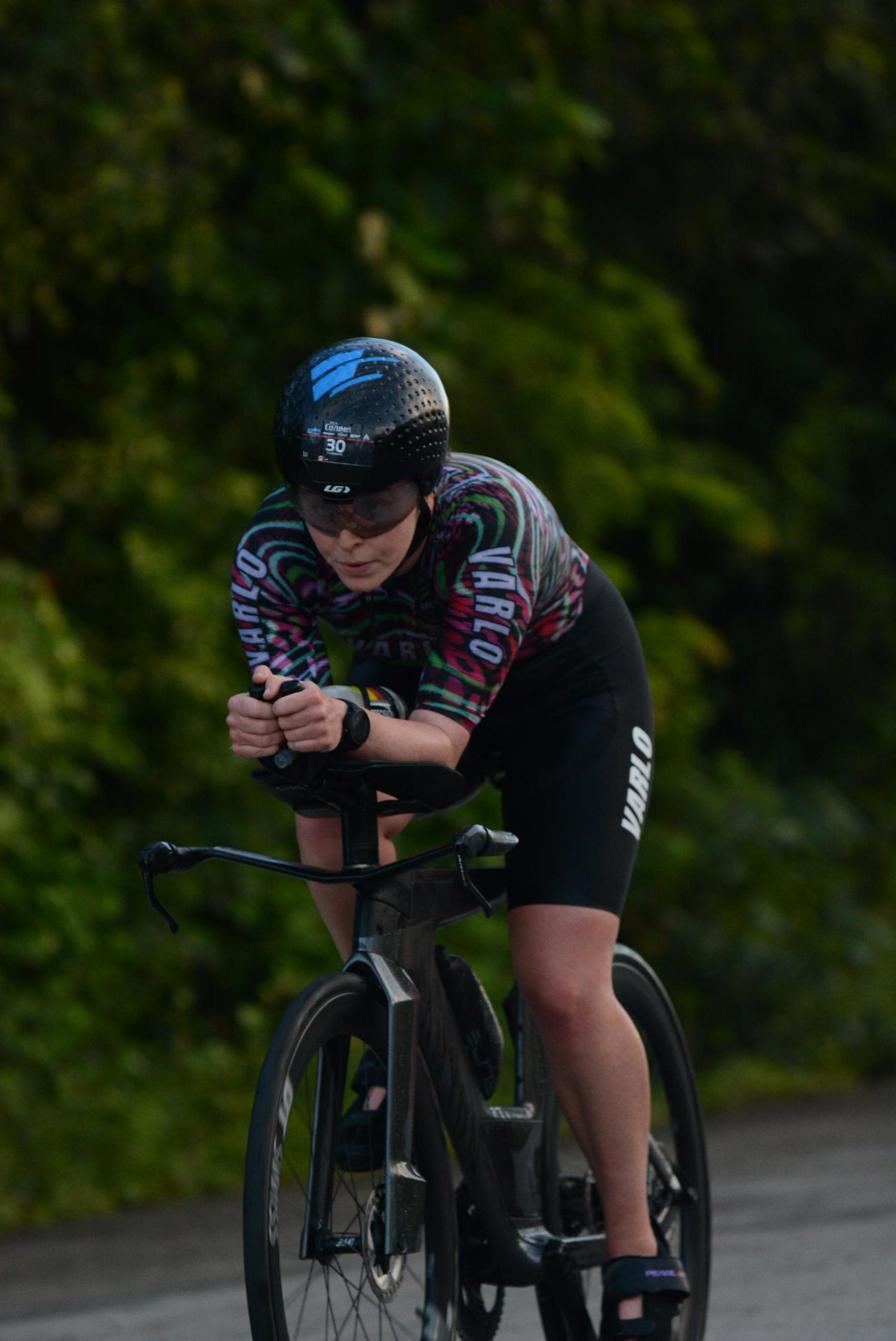 Close up of the athlete riding in an aero position on a triathlon bike. The entire background is green trees. 