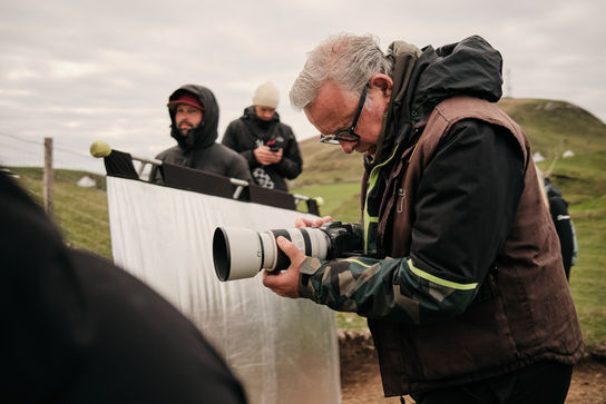 Man adjusting camera lens, reflector, and crew visible during outdoor shoot.