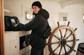 Man in black jacket using laptop, large wooden ship's wheel in cabin.
