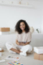 Smiling woman sits cross-legged on carpet, holding a tablet. Boxes and color palettes on floor. Minimalist white decor background.