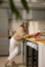 Older woman with grey hair reading a red book in a modern kitchen, leaning on a counter with cut pomegranates. Gray cabinets, cozy atmosphere.